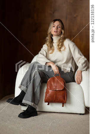 beautiful girl in gray pants and beige sweater posing with a small shiny brown leather backpack 122138136
