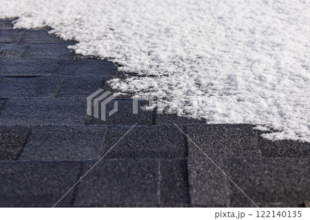 Snow-covered asphalt shingle roof with visible patterns and texture, illuminated by winter sunlight creating a striking contrast Snow-covered asphalt shingle roof with visible patterns and texture, illuminated by winter sunlight creating a striking contrast 122140135