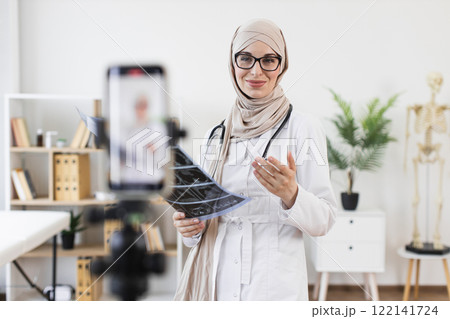 Muslim female doctor in hijab explaining x-ray results during online video lesson about treatment, showcasing modern medical education methods. 122141724