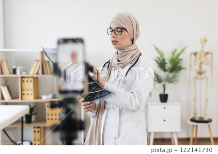 Muslim woman doctor recording video on phone explaining x-ray treatment. Young adult providing online medical education in modern office setting. 122141730