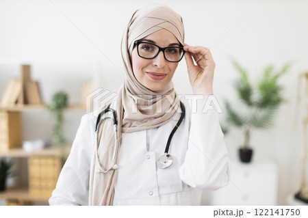Portrait of confident Muslim adult woman doctor wearing hijab with stethoscope. Middle Eastern female medical professional in modern office, exuding professionalism and compassion. Portrait of confident Muslim adult woman doctor wearing hijab with stethoscope. Middle Eastern female medical professional in modern office, exuding professionalism and compassion. 122141750