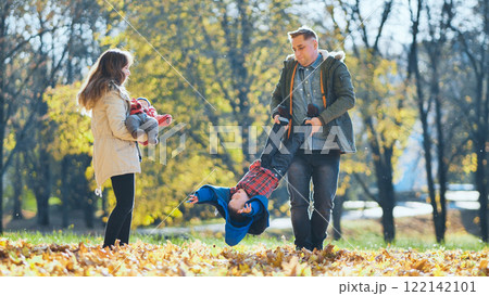 Happy family enjoying a sunny autumn day in a park, father holding his son upside down while mother holding baby, surrounded by colorful fallen leaves 122142101