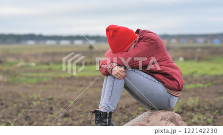 Lonely teenager wearing red beanie, crouching on rocky ground, concealing face between knees amid grassy landscape Lonely teenager wearing red beanie, crouching on rocky ground, concealing face between knees amid grassy landscape 122142230