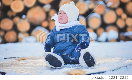 Tearful infant bundled in winter attire sobbing on snow covered wooden logs during wintry weather 122142233