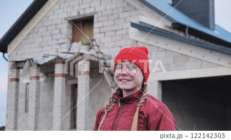 Confident young woman architect wearing a red beanie is smiling near a building under construction Confident young woman architect wearing a red beanie is smiling near a building under construction 122142303