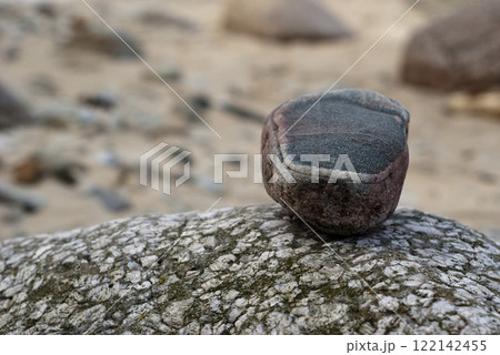Vibrant multicolored stone resting on weathered rock surface, set against blurred sandy beach backdrop for natural coastal vibe Vibrant multicolored stone resting on weathered rock surface, set against blurred sandy beach backdrop for natural coastal vibe 122142455