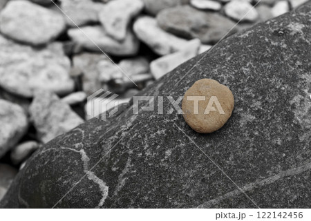 Simple composition featuring single light brown pebble resting on textured dark stone with blurred background of scattered rocks 122142456