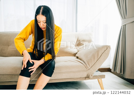 A woman seated on a sofa holds a painful knee displaying chronic tendon arthritis. Depicting Health Care and Medical Concept highlighting tendon inflammation and pain. 122144032