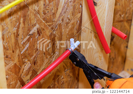 Contractor uses pliers to secure plumbing fitting on red piping in wood framed construction area. 122144714
