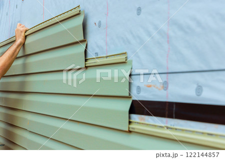 Construction worker carefully lays green siding on building exterior while ensuring proper fit. 122144857