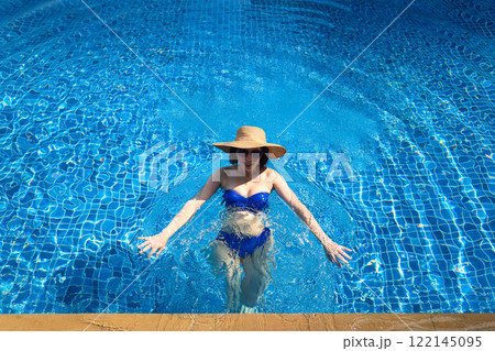 Blissful Summer Escape: A Woman in a Straw Hat and Sunglasses Joyfully Swimming in a Private Pool, Embracing the Relaxation and Serenity of a Sun-Kissed Day Under the Clear Blue Sky 122145095