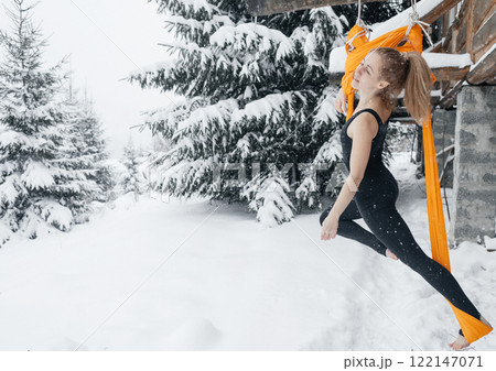 Young woman doing fly yoga in snowy forest in mountains. Yoga in hammock in winter against snow and fir trees. Gymnastic tricks outdoors in extreme conditions Young woman doing fly yoga in snowy forest in mountains. Yoga in hammock in winter against snow and fir trees. Gymnastic tricks outdoors in extreme conditions 122147071