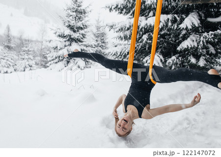 Young woman doing fly yoga in snowy forest in mountains. Yoga in hammock in winter against snow and fir trees. Gymnastic tricks outdoors in extreme conditions 122147072