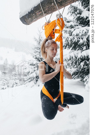 Young woman doing fly yoga in snowy forest in mountains. Yoga in hammock in winter against snow and fir trees. Gymnastic tricks outdoors in extreme conditions Young woman doing fly yoga in snowy forest in mountains. Yoga in hammock in winter against snow and fir trees. Gymnastic tricks outdoors in extreme conditions 122147076