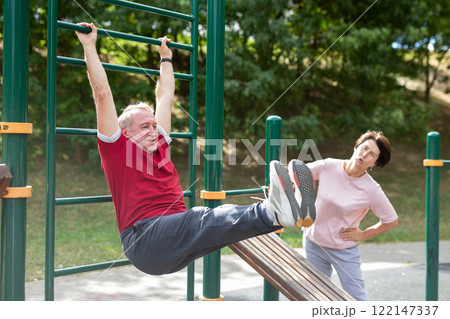 Elderly man and woman doing exercises on horizontal bars at outdoor sports ground Elderly man and woman doing exercises on horizontal bars at outdoor sports ground 122147337