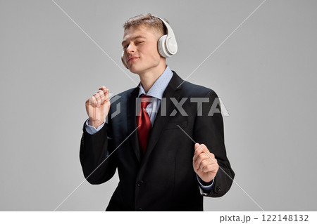 Overjoyed, relaxed young man, dressed formal attire, dancing while listening music in headphones against grey studio background. Overjoyed, relaxed young man, dressed formal attire, dancing while listening music in headphones against grey studio background. 122148132