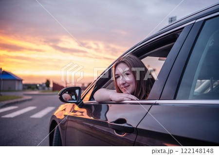 Happy young woman leaning out of car window, enjoying beautiful sunset view during road trip Happy young woman leaning out of car window, enjoying beautiful sunset view during road trip 122148148