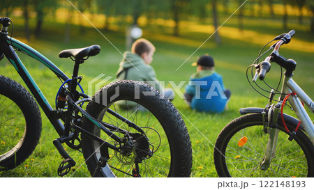 Boys resting on grass near parked bicycles, relaxing under bright sunlight after cycling through park landscape Boys resting on grass near parked bicycles, relaxing under bright sunlight after cycling through park landscape 122148193