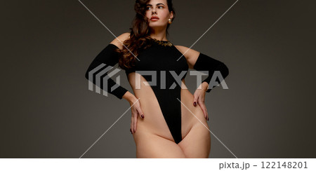 Plus size woman standing tall in black bodysuit, holding hands on hips, radiating strength, confidence, and natural elegance against grey studio background. 122148201