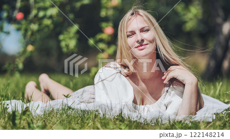 Blonde woman relaxing on soft blanket, resting amid lush grass and fruit trees under warm summer sunlight Blonde woman relaxing on soft blanket, resting amid lush grass and fruit trees under warm summer sunlight 122148214