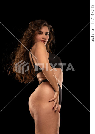 Side view photo of stylish and confident plus-size woman, wearing black lingerie, posing looking at camera over shoulder against black studio background. Side view photo of stylish and confident plus-size woman, wearing black lingerie, posing looking at camera over shoulder against black studio background. 122148215