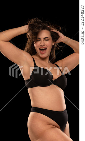 Happy, joyful young woman, plus size woman posing, showing body positivity and self-acceptance against black studio background. Happy, joyful young woman, plus size woman posing, showing body positivity and self-acceptance against black studio background. 122148227