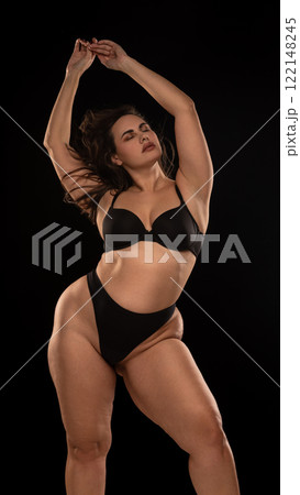 Overweight young woman in black underwear, posing in relaxed posture with closed eyes and holding arms raised against black studio background. 122148245