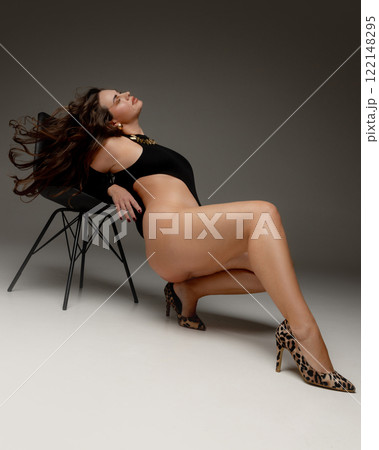 Woman, model plus size, leaning back on chair, her hair flowing freely, celebrating empowerment and femininity against grey studio background. 122148295