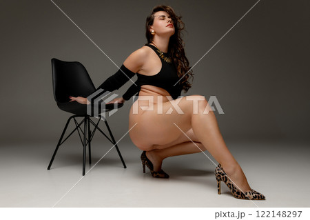Woman, plus size model, leaning against chair, dressed black bodysuit and leopard heels, shows confidence and poise against grey studio background. Woman, plus size model, leaning against chair, dressed black bodysuit and leopard heels, shows confidence and poise against grey studio background. 122148297