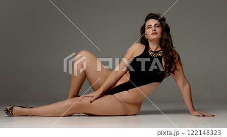 Horizontal photo of young brunette woman, dressed stylish black body, lying on floor against grey studio background, posing showing her curvy body 122148323