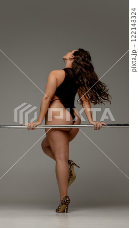 Back view photo of curvy woman, dressed black body and heels with animal print, leaning elegantly on barre against grey studio background. Back view photo of curvy woman, dressed black body and heels with animal print, leaning elegantly on barre against grey studio background. 122148324