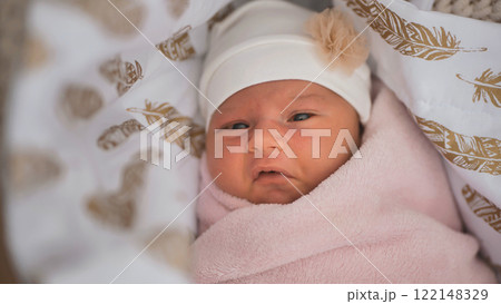 Newborn baby girl wearing white cap and wrapped in pink blanket is lying in crib with feather pattern bedding Newborn baby girl wearing white cap and wrapped in pink blanket is lying in crib with feather pattern bedding 122148329