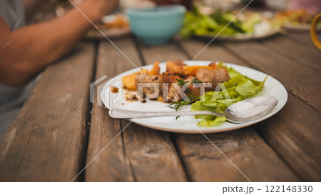 Leftovers of sausages, potatoes and salad on white plate with spoon on wooden table after eating outside Leftovers of sausages, potatoes and salad on white plate with spoon on wooden table after eating outside 122148330