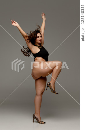 Beautiful chubby woman posing dressed black body and raising hands against grey studio background, celebrating charm of soft curves. Side view. 122148333