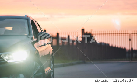 Sleek vehicle parked roadside, gleaming headlights illuminating dramatic sunset sky with vibrant orange pink gradient 122148337