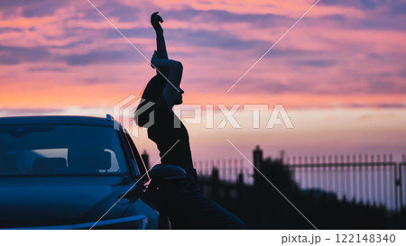 Silhouette of a young woman dancing and enjoying the sunset near her car, expressing freedom and a carefree attitude 122148340