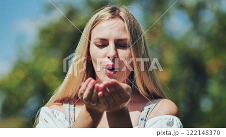 Blonde woman softly blowing kiss, lush green background, sunlight warming her face 122148370