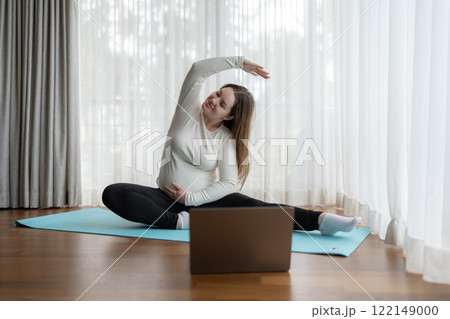 Young pregnant woman practicing yoga at home with laptop. Expectant mother doing prenatal training class. Young pregnant woman practicing yoga at home with laptop. Expectant mother doing prenatal training class. 122149000