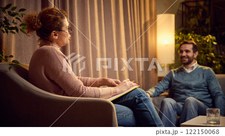 Female psychologist with notes explaining something to a male client seated on a couch in a warm, cozy therapy space. Female psychologist with notes explaining something to a male client seated on a couch in a warm, cozy therapy space. 122150068