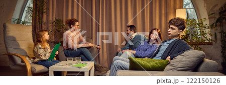 Family of four, including parents, teen boy and school-age girl visiting psychologist, sitting in warmly-lit room, discussing problems and finding solutions 122150126