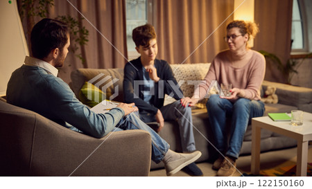 Psychologist leads therapy session with mother and her teenage son, discussing family issues in calm environment. 122150160