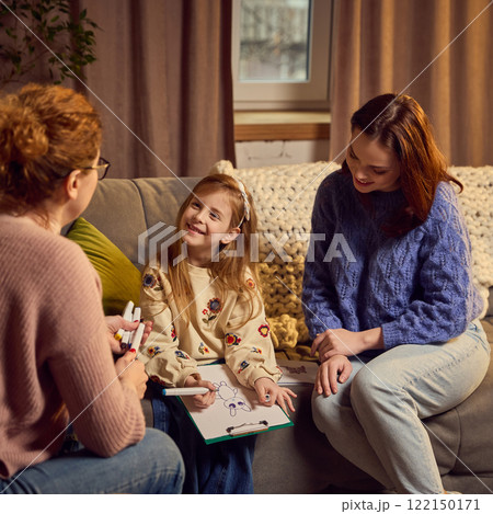 Young girl shares joyful moment with psychologist while drawing, her smiling mother observes drawing warmly, creating supportive therapy setting. 122150171