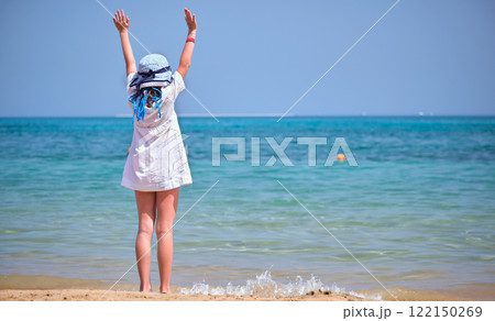 Rear view of child girl in white dress and hat standing barefoot with arms raised up on sandy beach enjoying tropical vacations looking at blue sea water 122150269