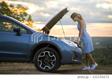 Puzzled young female driver standing near a broken car with open up hood inspecting her vehicle motor. 122150272