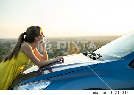 Happy young woman driver in yellow dress enjoying warm summer evening standing beside her car. Travelling and vacation concept. Happy young woman driver in yellow dress enjoying warm summer evening standing beside her car. Travelling and vacation concept. 122150372