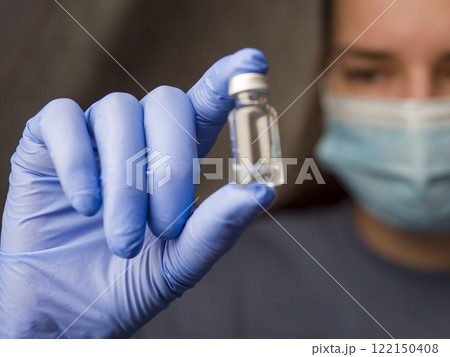 Medical professional examining clear liquid vial, wearing blue latex gloves and surgical mask in clinical setting 122150408