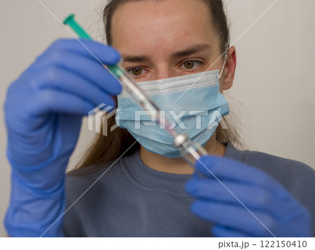 Female doctor wearing blue nitrile gloves and surgical mask, carefully preparing vaccine by filling syringe from vial during the ongoing covid-19 pandemic 122150410