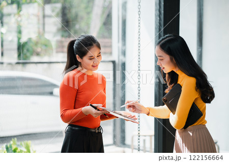 Two women in a bright office discussing work, using a digital tablet, dressed in vibrant colors. 122156664