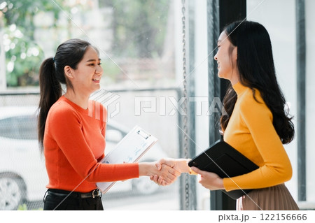 Two women in a modern office setting, shaking hands and smiling, symbolizing successful business collaboration. 122156666