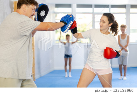 Woman in boxing gloves punching pads in hands of husband during family traning Woman in boxing gloves punching pads in hands of husband during family traning 122158960